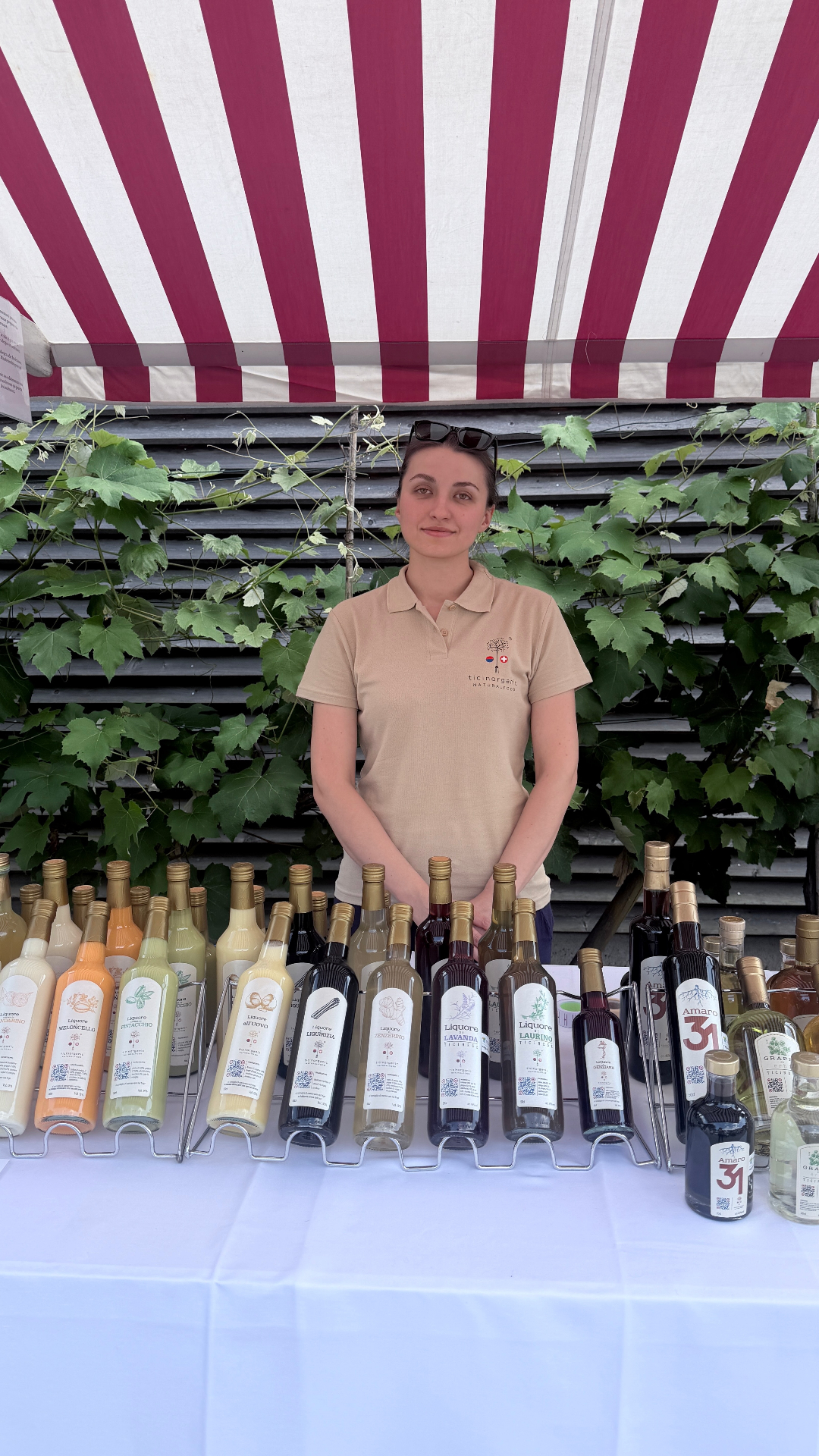 Banco del mercato Ticinorganic con bottiglie di liquori artigianali e operatrice in polo brandizzata